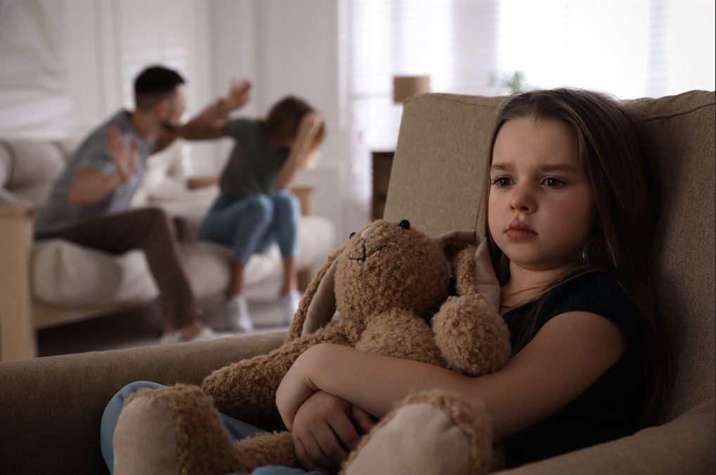 Young girl hugs a teddy bear while listening to parents fighting in the background